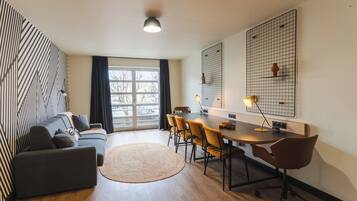 Una sala de estar moderna con un sofá, una mesa de comedor con sillas, un ventanal grande y una lámpara fijada a la pared.