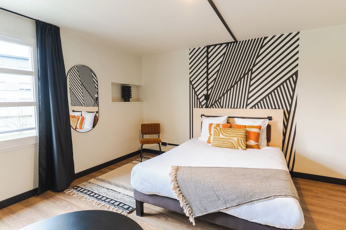 A modern bedroom with a large bed, a side table, a chair, a mirror, and a geometric-patterned headboard.
