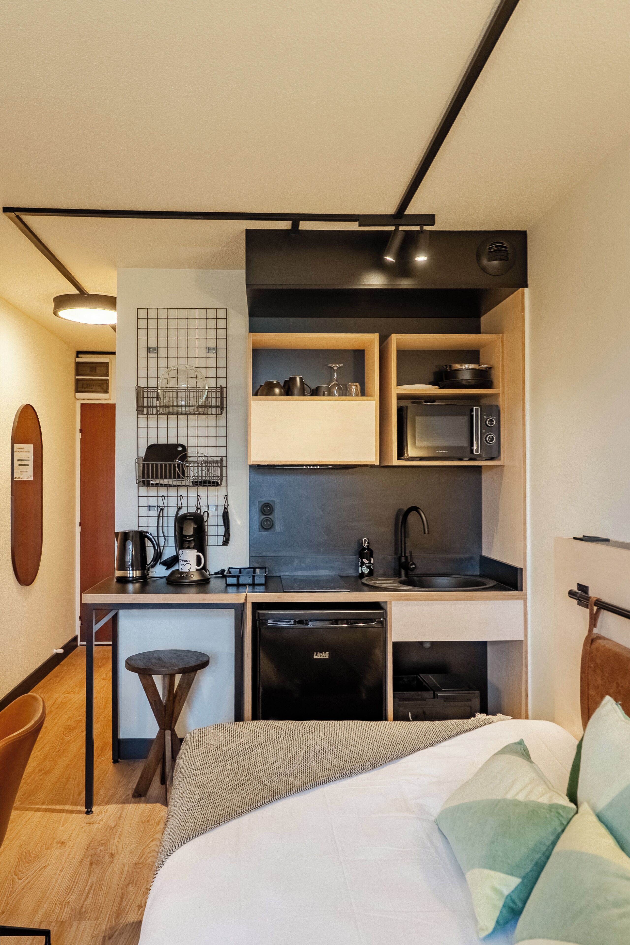 A compact hotel room with a kitchenette, a bed with green pillows, a small round table, and a wooden floor.