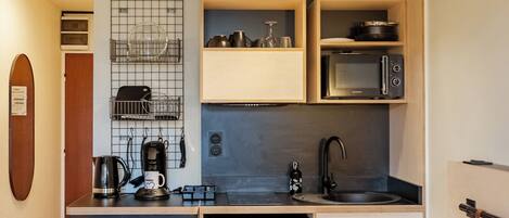 A compact hotel room with a kitchenette, a bed with green pillows, a small round table, and a wooden floor.