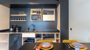 A compact kitchen with a black countertop, a sink, a coffee maker, and a refrigerator. There are wooden shelves above the countertop holding kitchenware. - Cowool Compiègne (Compiègne)