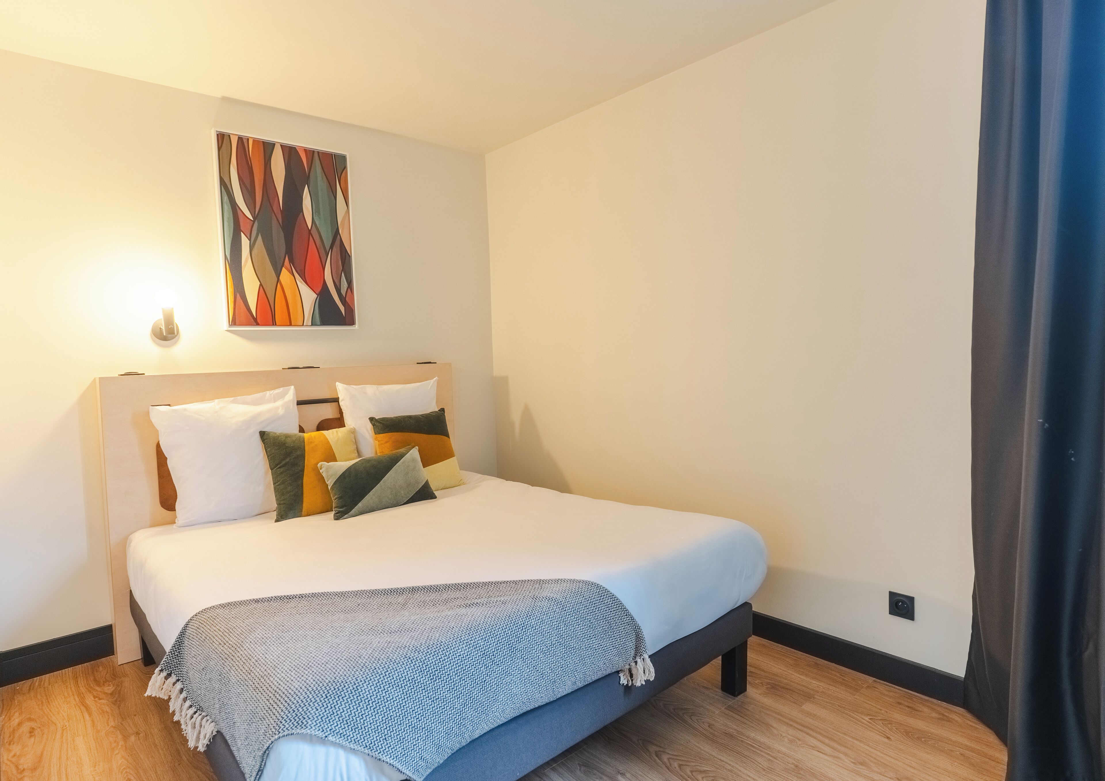 Una cama bien hecha con edredón blanco y varios almohadones, una lámpara de pared y un cuadro abstracto colorido sobre la cama.