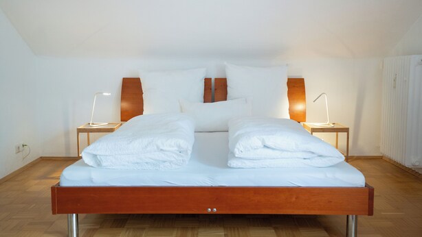 A modern bedroom with a wooden bed frame, white linens, two bedside lamps, and a wooden headboard. - Guesthouse Elena Apartment 23 (Kassel)