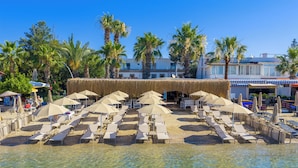 Un complexe hôtelier en bord de mer, doté de chaises longues et de parasols, de palmiers et d’un bâtiment au toit de chaume.