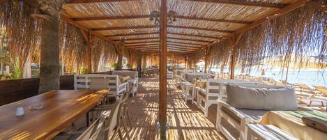 An outdoor seating area with wooden tables and white cushioned benches, a thatched roof, and a view of the water.