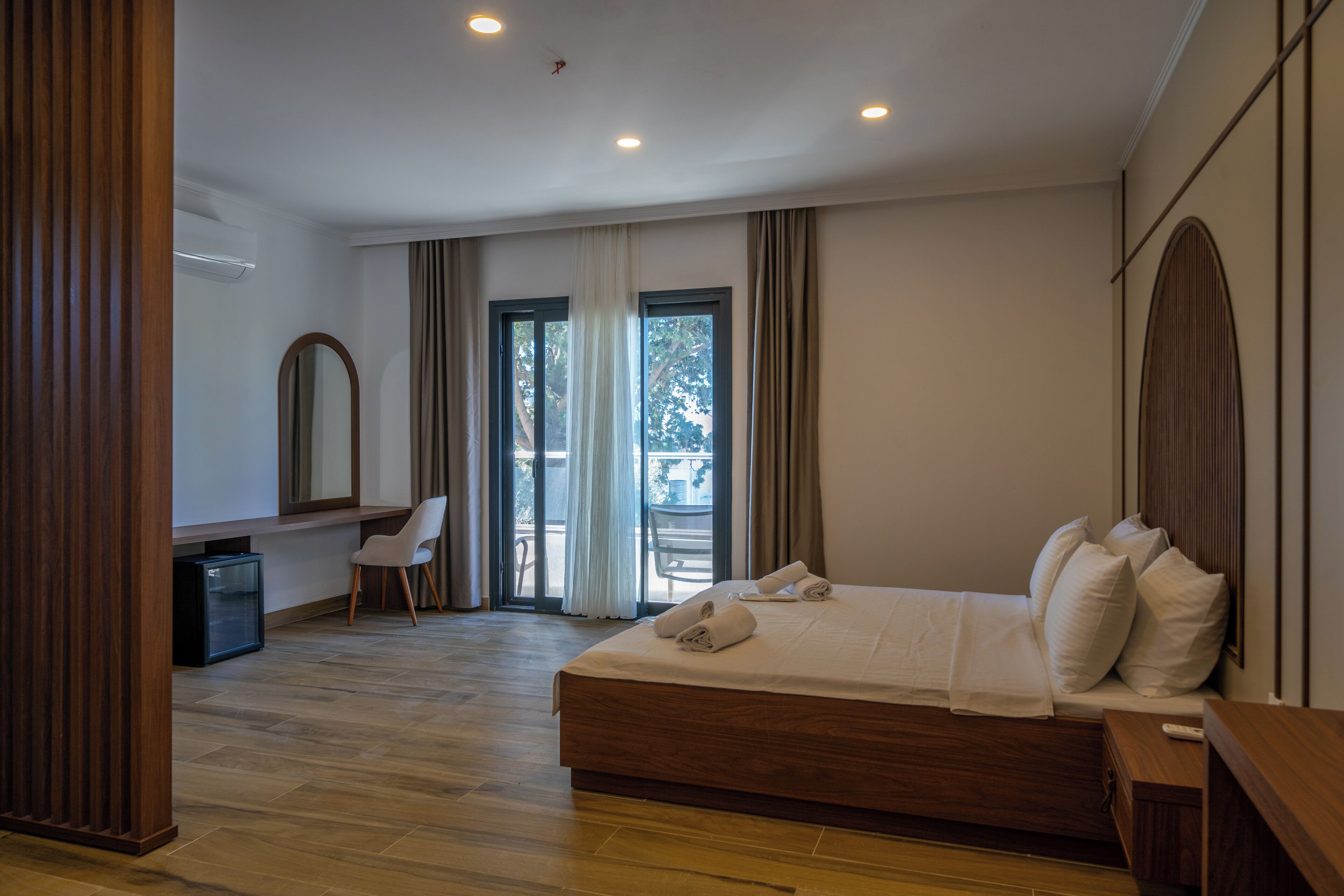 Habitación de hotel moderna con piso de madera, una cama grande con ropa blanca, un escritorio con silla y un balcón con vista a árboles.