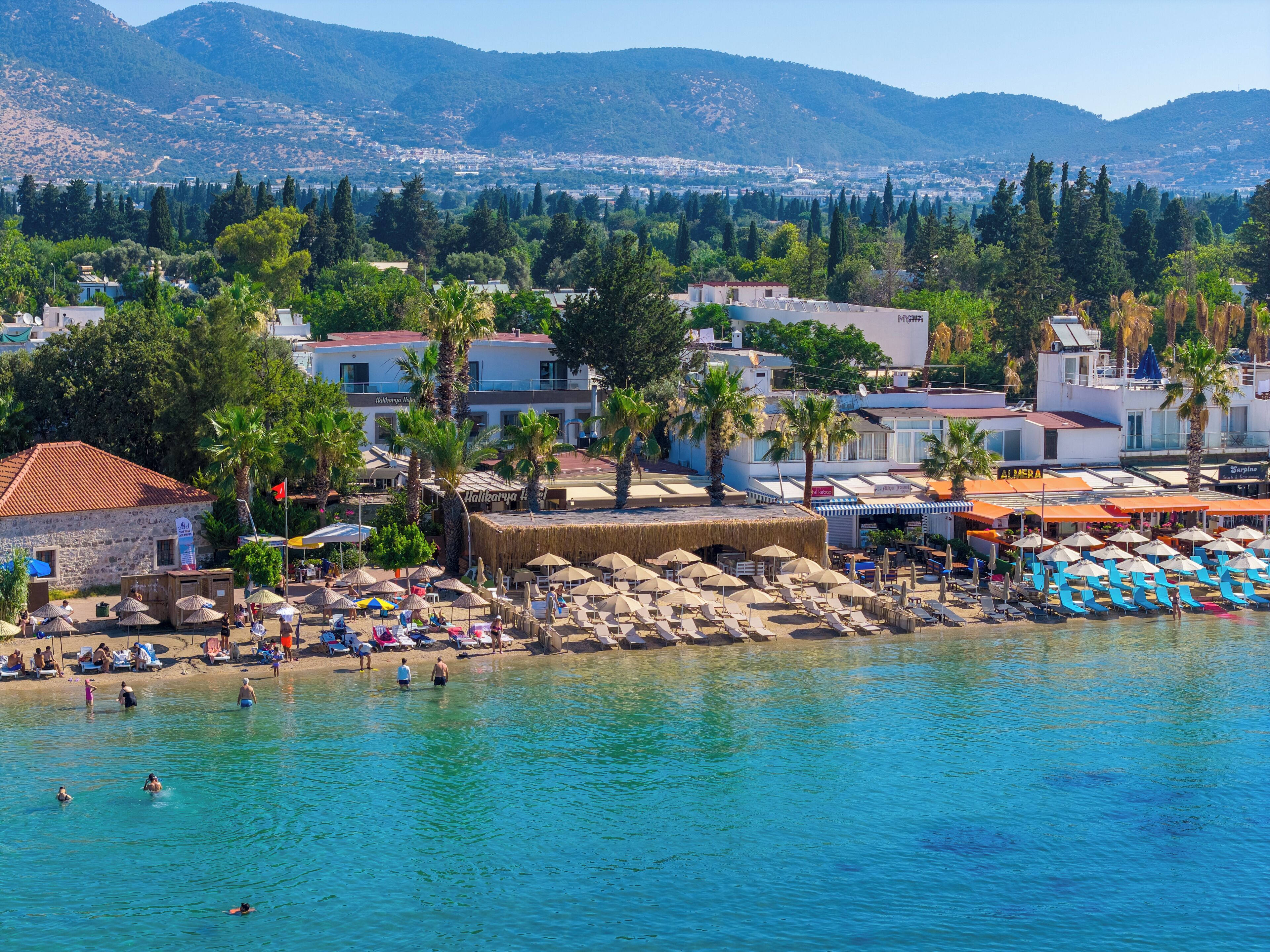 A coastal resort with beach umbrellas, swimming pools, and a view of the sea and mountains.