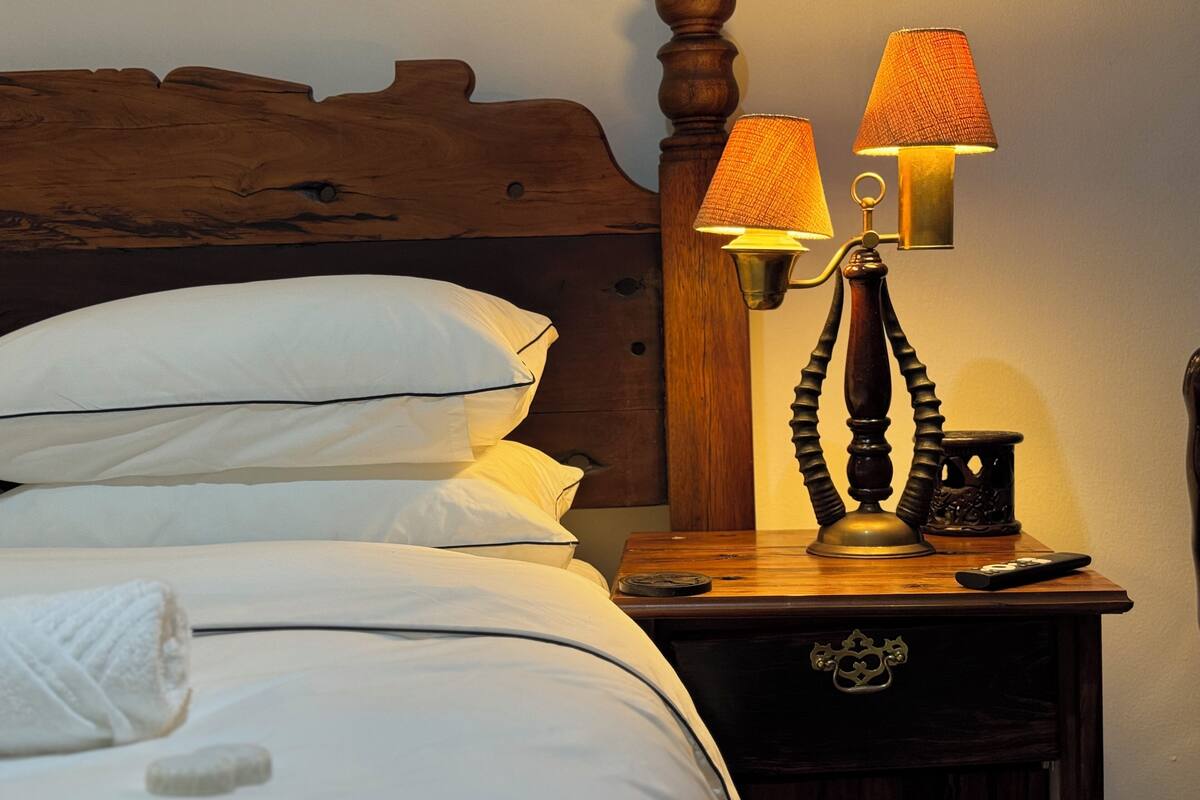 A neatly made bed with white linens and pillows, a wooden headboard, a bedside table with a lamp, a remote control, and a small decorative item.