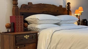 Uma cama bem arrumada com lençóis brancos, cabeceira de madeira, mesa de cabeceira com um globo e livros, e uma luminária ao lado.