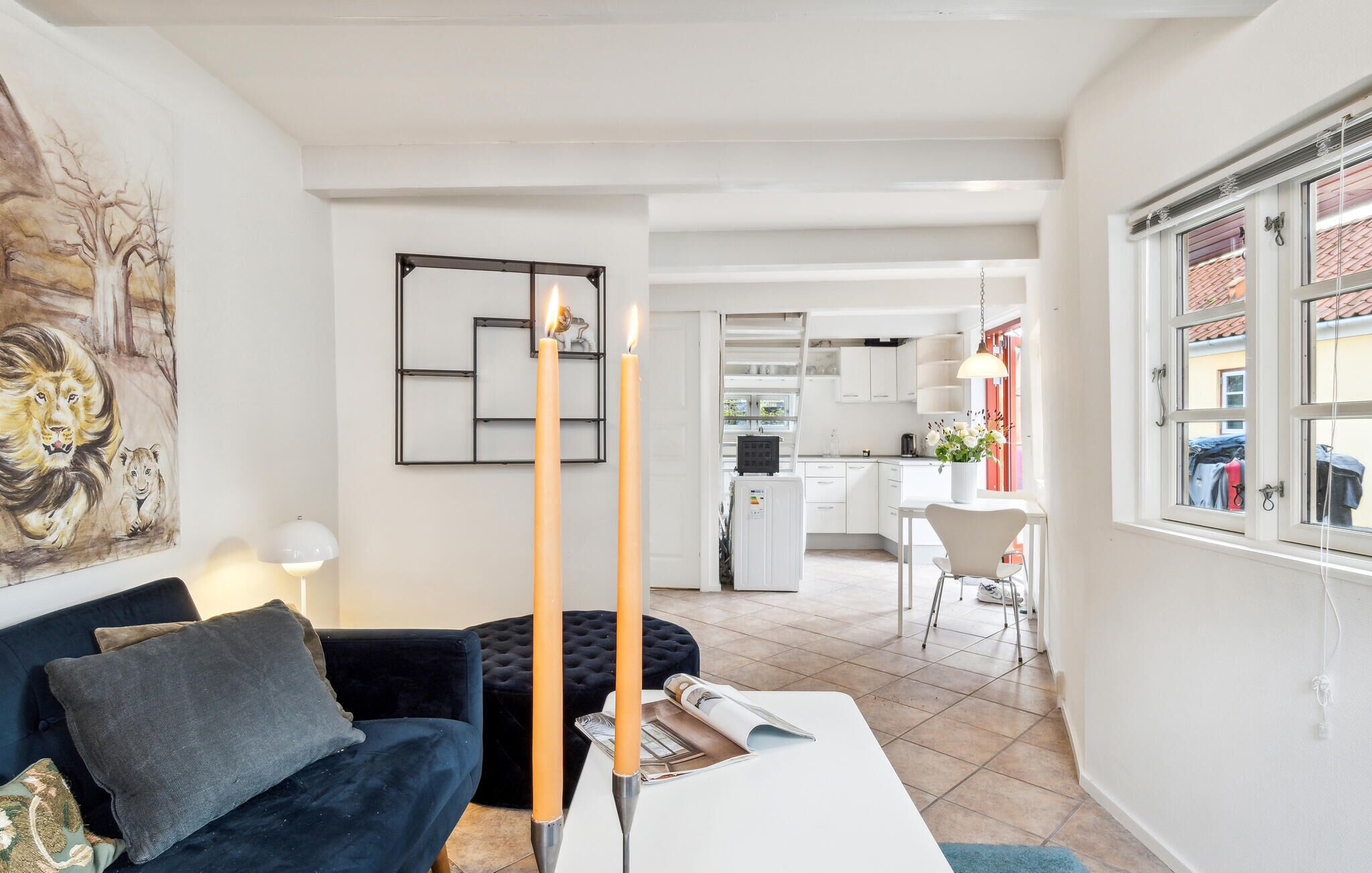 Ein modernes Wohnzimmer mit einem dunkelblauen Sofa, einem weißen Couchtisch und einem Gemälde mit Löwen. In der Mitte steht eine hohe Kerze, und im Hintergrund ist eine Küchenzeile erkennbar.
