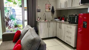 A compact kitchen with a sink, stove, and microwave. A grey sofa with red cushions is in the foreground.