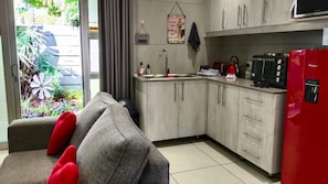 Une cuisine compacte équipée d’un évier, d’une cuisinière et d’un four à micro-ondes. Un canapé gris avec des coussins rouges se trouve au premier plan.
