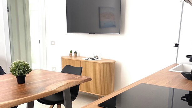 A modern conference room with a wooden table, black chairs, a small potted plant, a wooden cabinet, and a flat-screen TV mounted on the wall. - Domus Luxi - Teulada Design Apartment | Style and Comfort (Teulada)