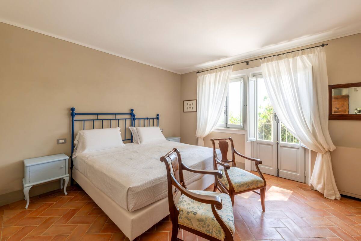 Une chambre à coucher avec un lit, deux tables de chevet, une chaise en bois, une fenêtre avec des rideaux et un miroir au mur.