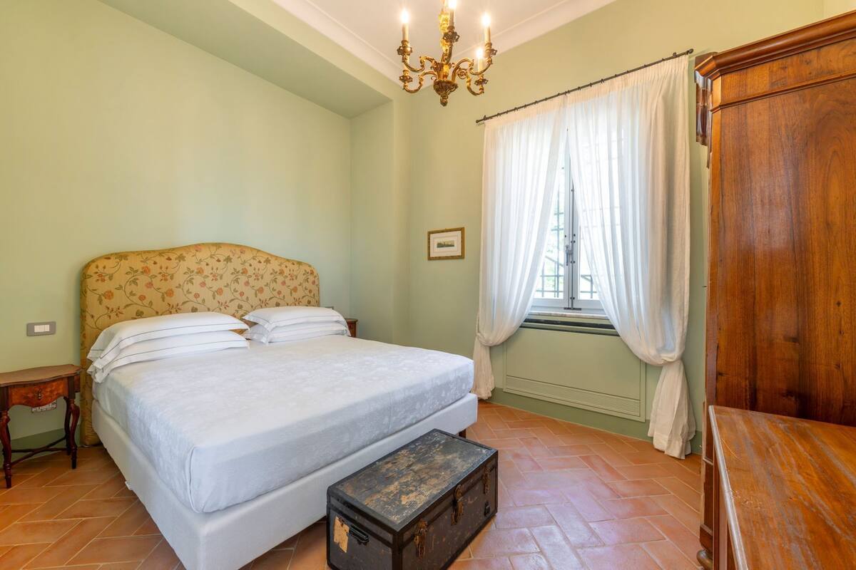 Une chambre à coucher comprenant un lit, une commode en bois, une table de chevet, une fenêtre avec des rideaux et un lustre.