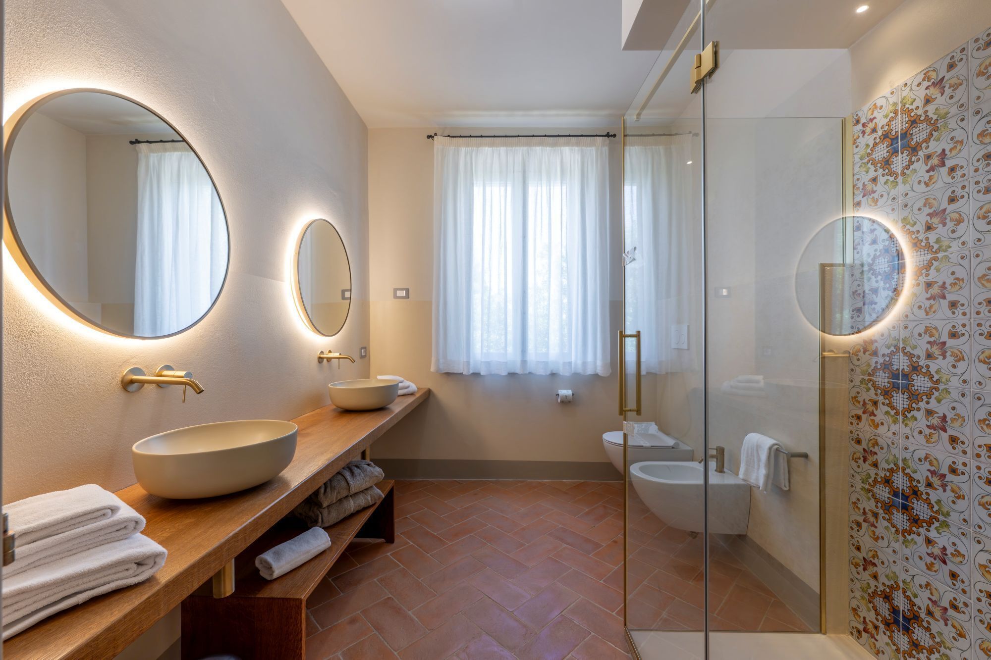 A modern bathroom with a large oval mirror, a wooden vanity, a freestanding bathtub, and a glass-enclosed shower.