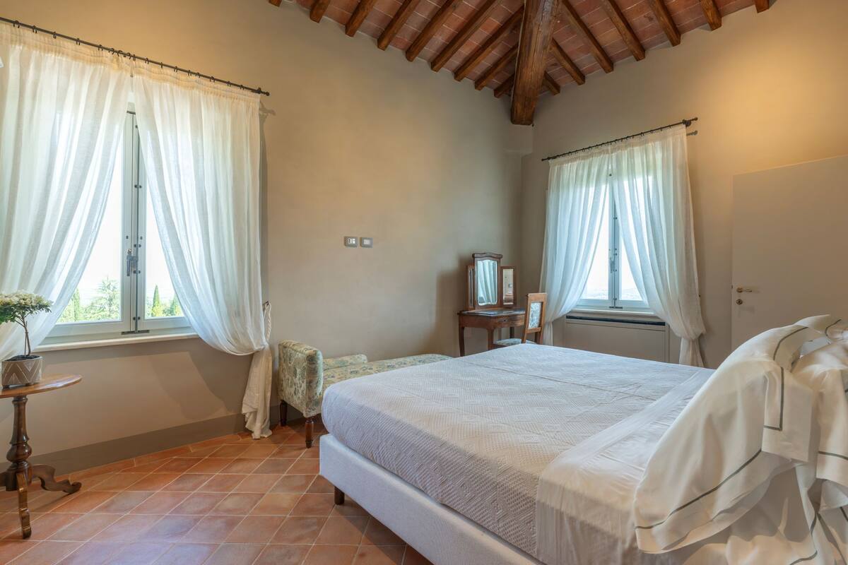 Une chambre à coucher avec un lit, une petite table sur laquelle est posée une plante, un miroir et une fenêtre avec des rideaux.