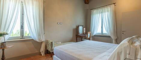 A bedroom with a bed, a small table with a plant, a mirror, and a window with curtains.