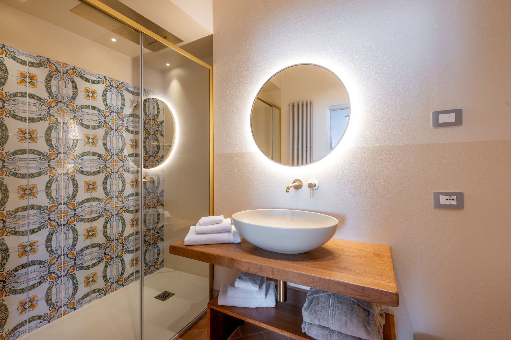 A bathroom with a large oval sink, a mirror with integrated lighting, and a shower with decorative tiles.