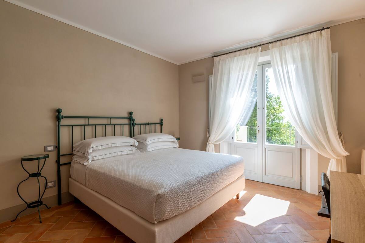 Une chambre à coucher avec un lit, une table de chevet, une fenêtre avec des rideaux et une vue sur la verdure à l’extérieur.