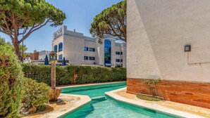 Une piscine à l’eau d’un bleu limpide, entourée de verdure et bordée d’un bâtiment à la façade blanche et aux fenêtres bleues.