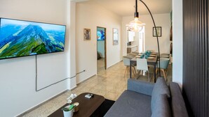 A modern living room with a large flat-screen TV, a sofa, a coffee table with a plant, and a dining area with a table and chairs.