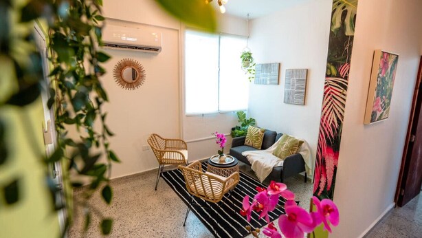A living room with a sofa, two wicker chairs, a coffee table, and a black and white striped rug. There is a window with a view of greenery outside, and a wall-mounted air conditioner.