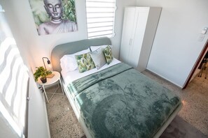 A bedroom with a bed, a nightstand, a potted plant, a wall-mounted light, and a window with blinds.