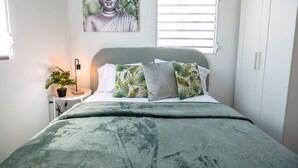 A bedroom with a bed, pillows, a nightstand, a lamp, a wall art of a Buddha, and a window with blinds.