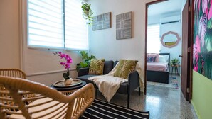 A cozy living room with a sofa, a wicker chair, a small table with a vase of flowers, and a bed in the background.