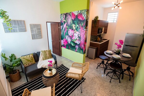 A compact living space with a sofa, a dining table, a kitchen area, and a large tropical-themed painting.