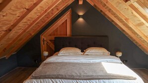 Une chambre avec un plafond en pente, des poutres en bois, une tête de lit sombre, deux lampes de chevet et un lit recouvert de linge de maison blanc.