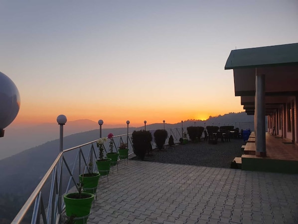 Une terrasse agrémentée de plantes en pot, offrant une vue sur le coucher de soleil au-dessus d’un paysage montagneux.
