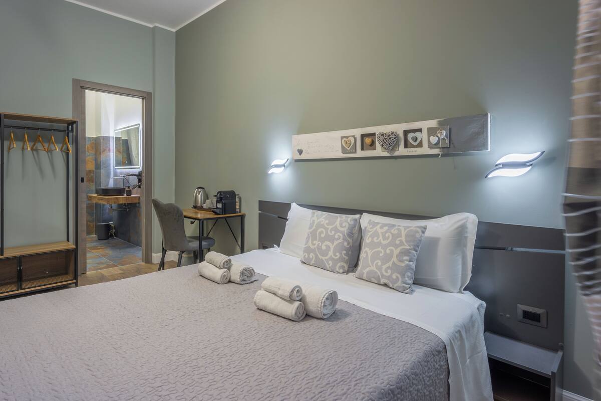 A neatly made bed with rolled towels, a desk with a chair, and a mirror in a room with light green walls and a shelf with decorative items.