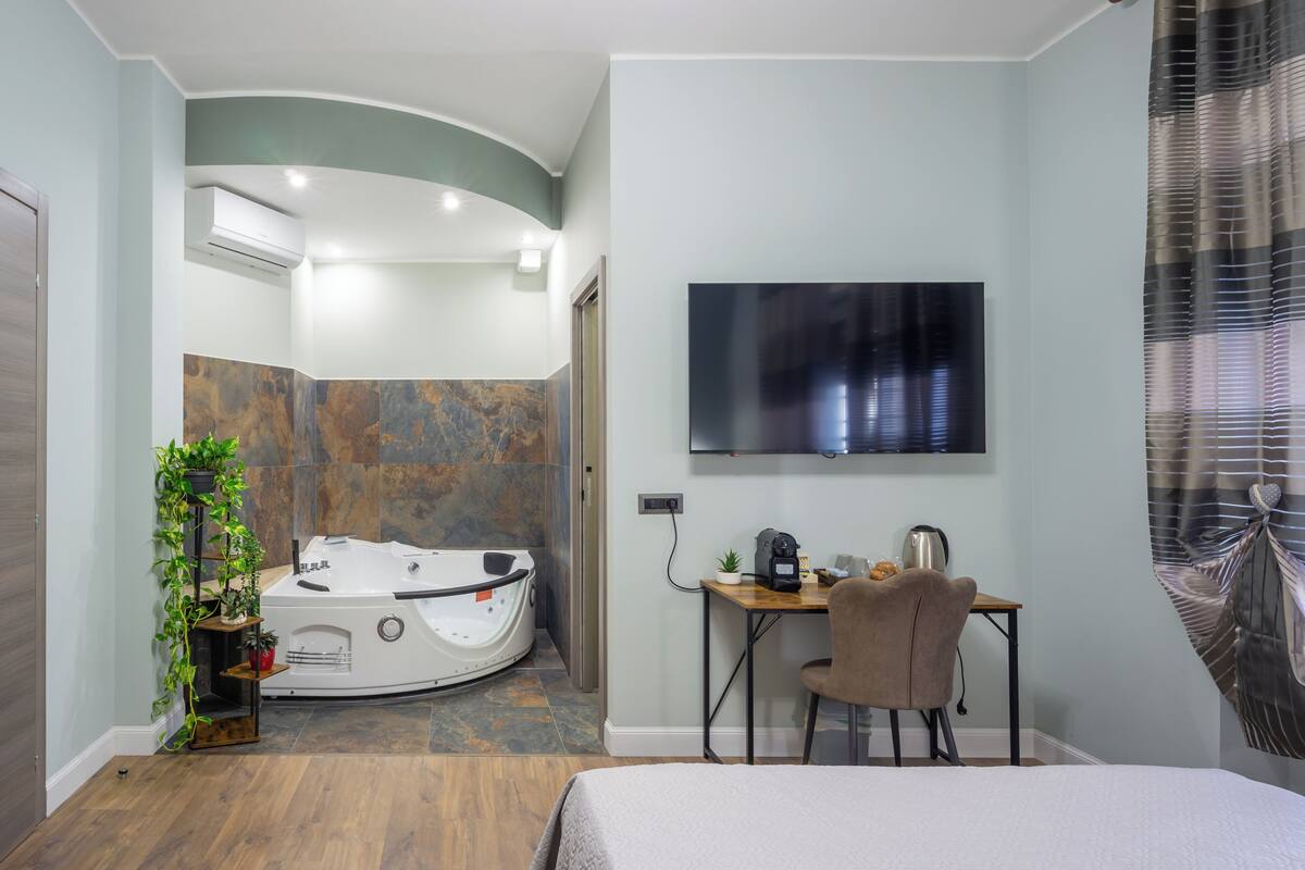 A modern hotel room with a curved ceiling, a large flat-screen TV mounted on the wall, a desk with a chair, a small potted plant, and a jacuzzi tub.