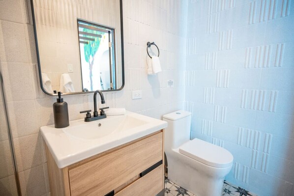 Un baño moderno con un lavamanos blanco, grifo negro y un espejo encima. Hay un inodoro con tapa blanca y una toalla colgada en la pared.