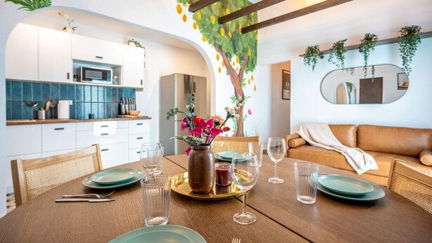 Un comedor con una mesa de madera preparada para cuatro personas, un florero con flores y una cocina al fondo.
