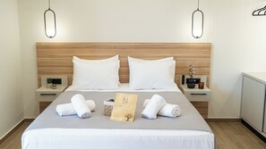 A neatly made bed with white linens, two white pillows, and two rolled towels on the bed. There is a small bedside table with a plant, a candle, and a card. The room has wooden paneling on the walls and a light fixture above the bed. - Holy Sea Smart Suites ( POSEIDON ) (Chania)