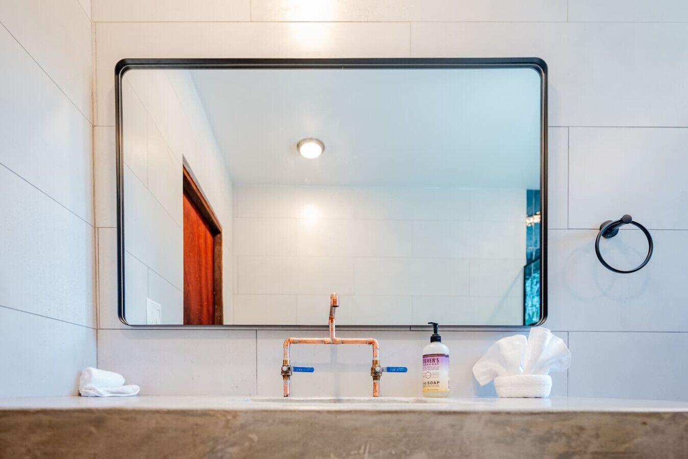 A bathroom with a large mirror, a sink with a copper faucet, a bottle of soap, and a towel on the counter.