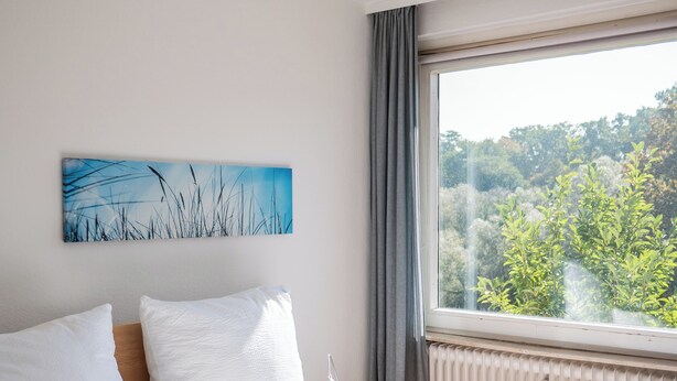 Ein ordentlich bezogenes Bett mit weißen Leinen, ein Fenster mit Blick auf Grünflächen und ein an der Wand hängendes Kunstwerk.