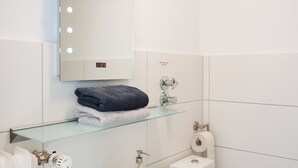 Ein modernes Badezimmer mit einem weißen Waschbecken, einer Glasablage mit Handtüchern, einer Toilette und einem beleuchteten Spiegel.