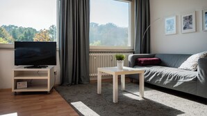 Ein modernes Wohnzimmer mit einem Flachbildfernseher auf einem weißen Standfuß, einem grauen Sofa, einem weißen Couchtisch mit Pflanze und einem großen Fenster mit Vorhängen.