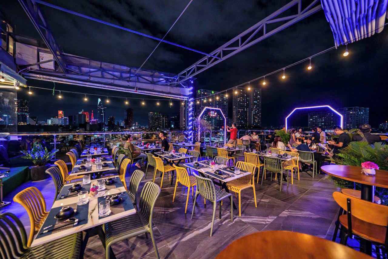 An outdoor dining area with tables and chairs, city skyline views, and string lights.