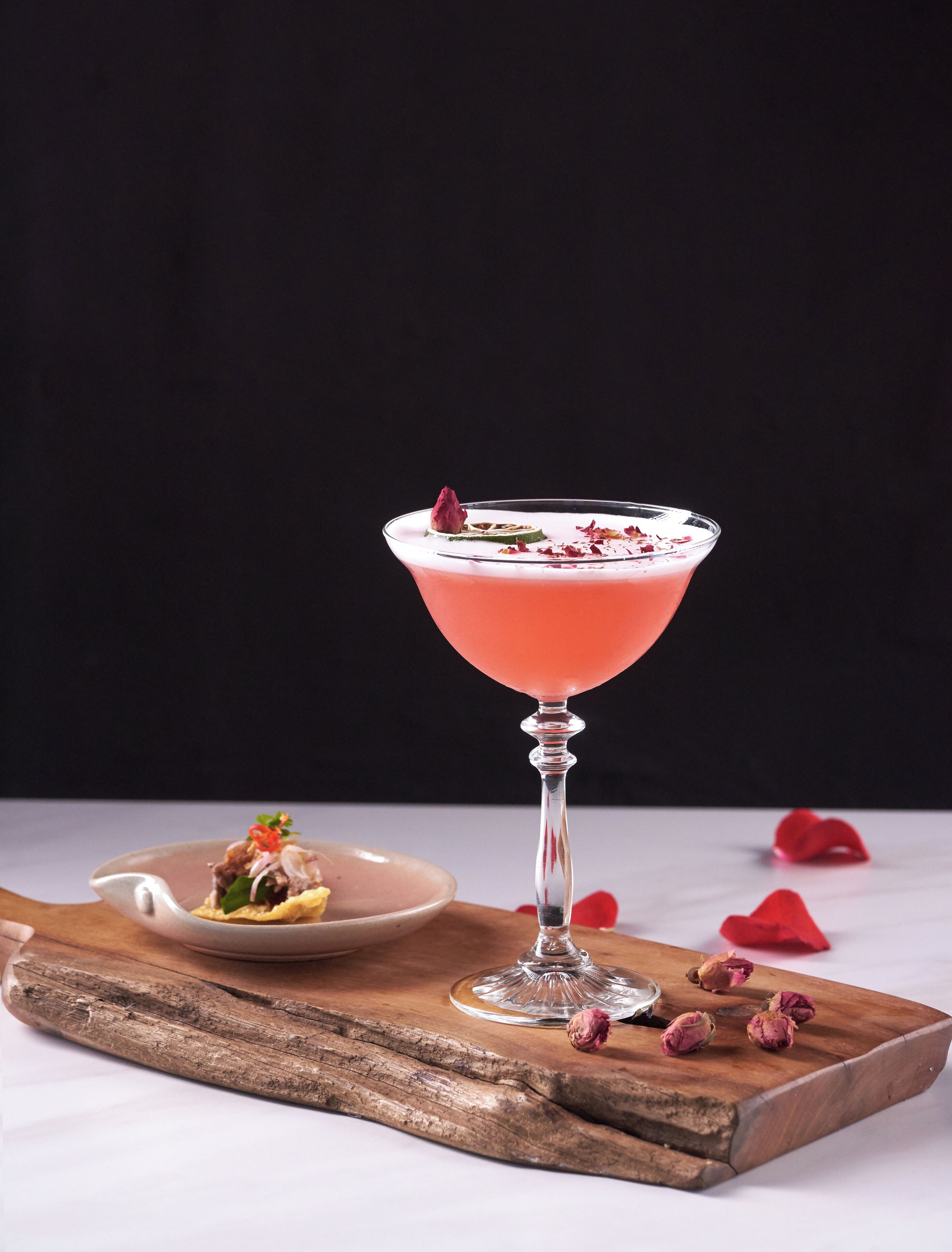 A cocktail in a martini glass with a rose garnish, placed on a wooden board with a small dish of food beside it.