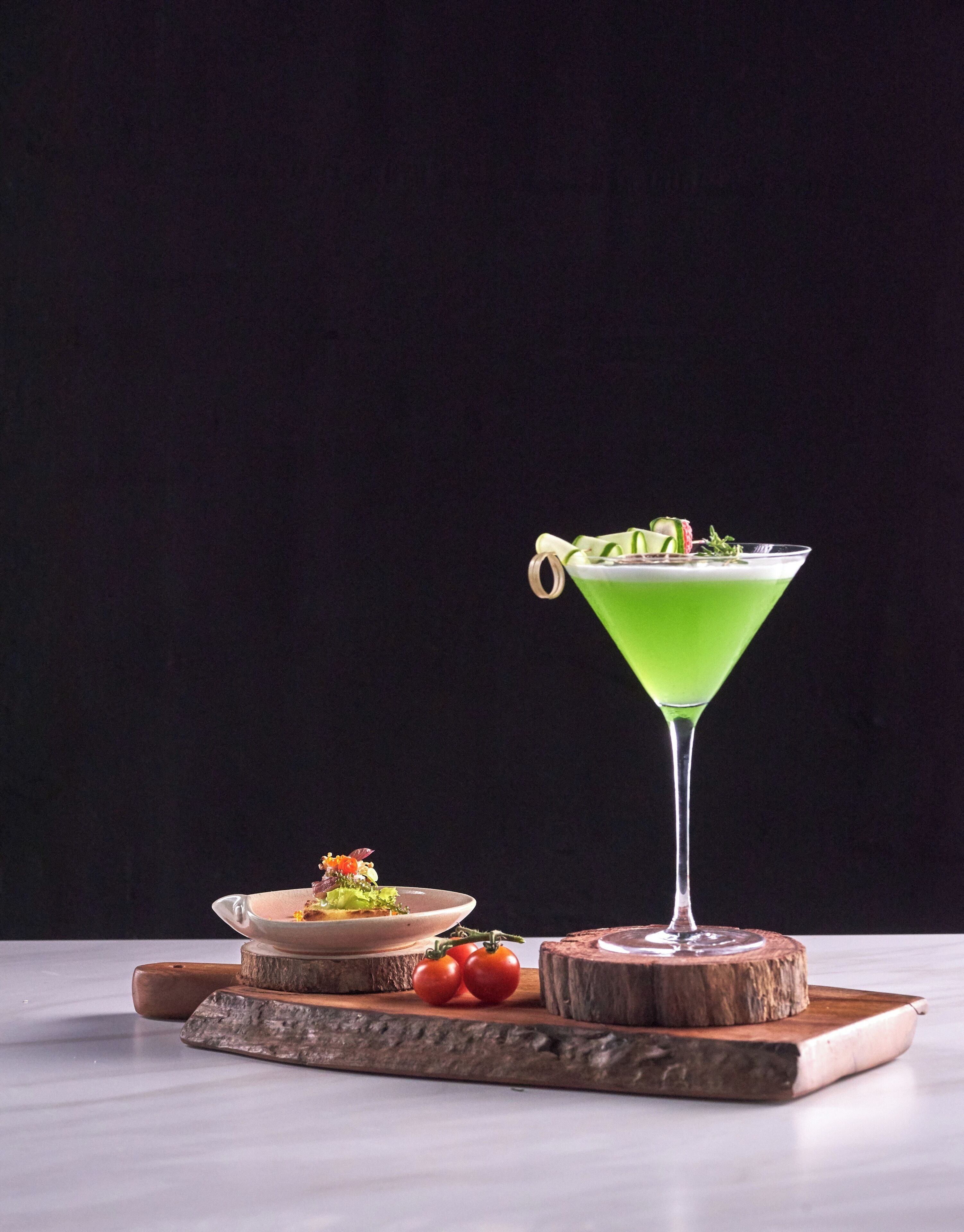 A martini glass with a green liquid and garnishes on a wooden coaster.
