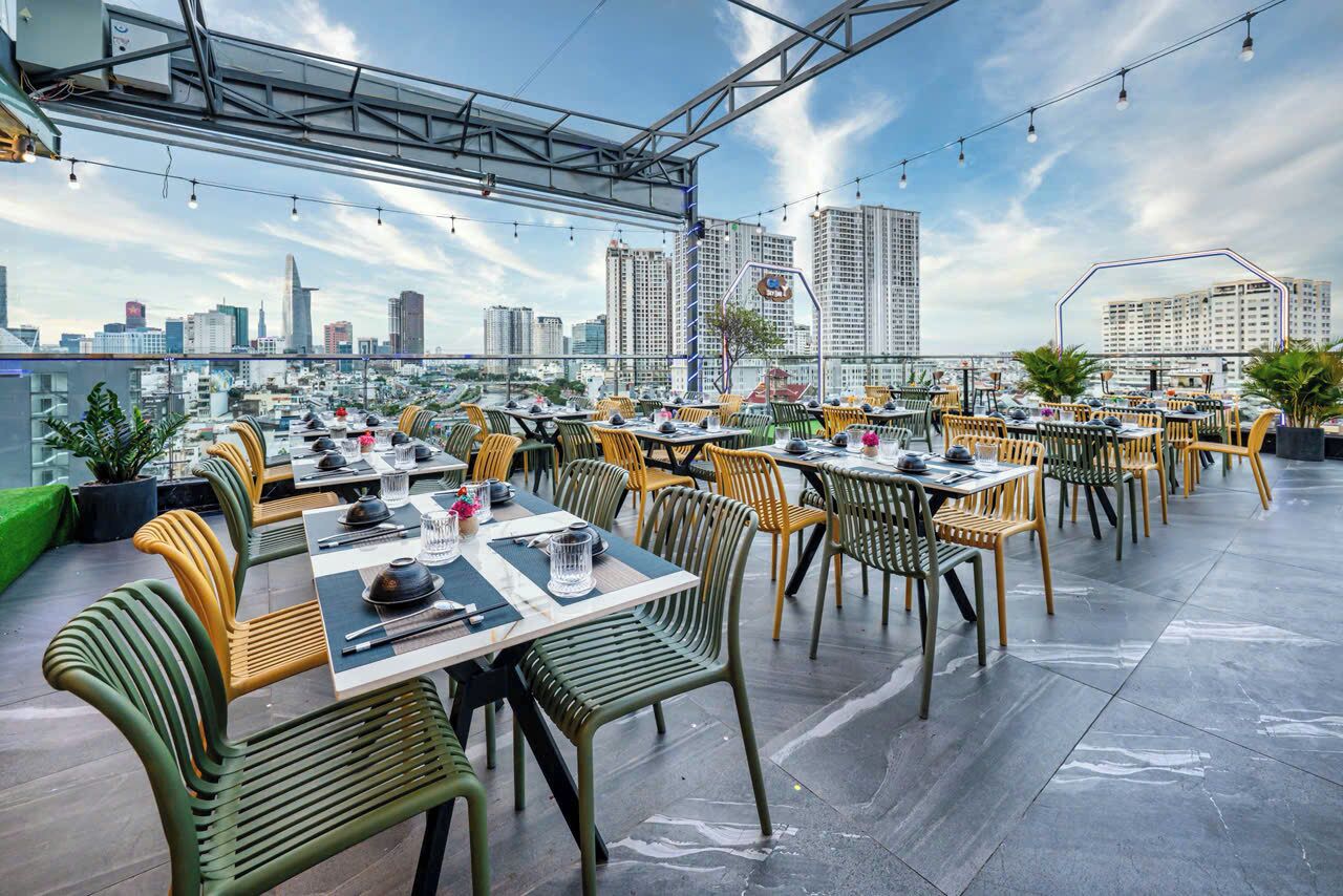 Un área de comedor al aire libre con varias mesas y sillas, un horizonte urbano al fondo y cielo despejado.