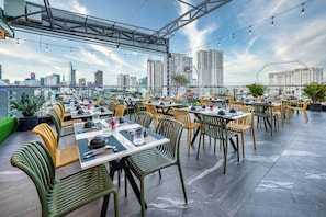 An outdoor dining area with multiple tables and chairs, a city skyline in the background, and a clear sky. - G8 Riverside Hotel & Rooftop Skybar (Ho Chi Minh City)