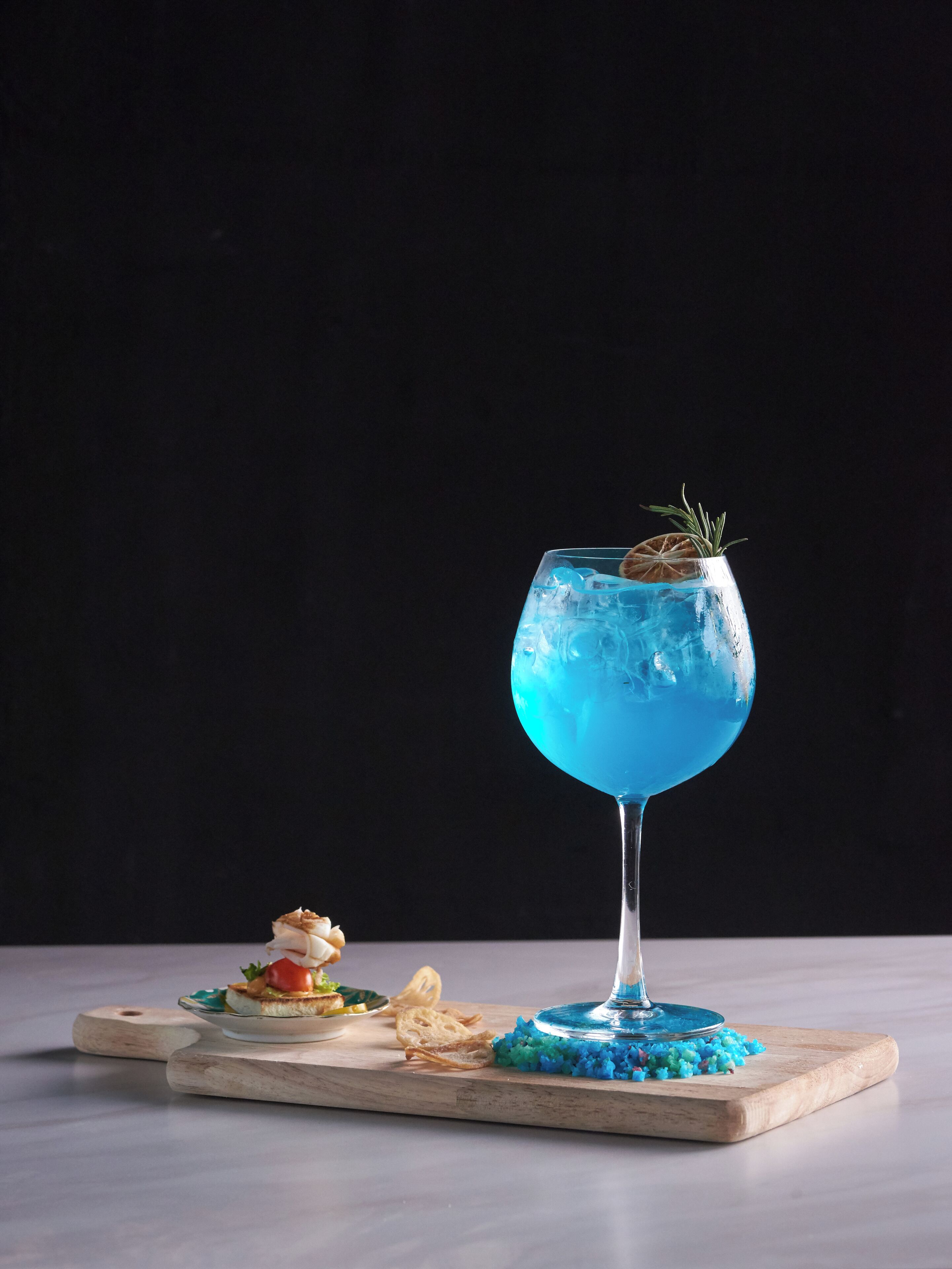 Un vaso de líquido azul con una ramita de romero sobre una tabla de madera.