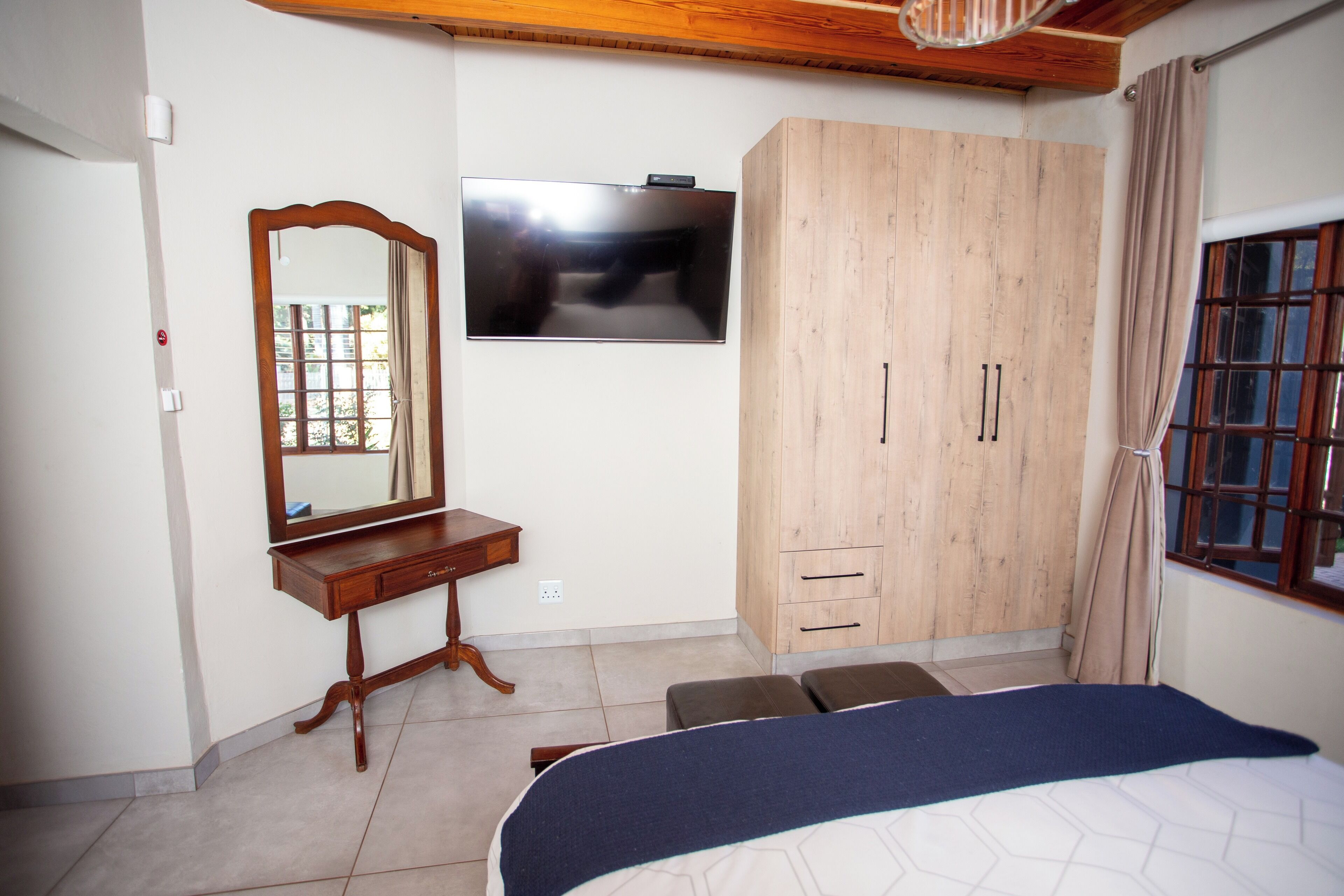 Une chambre à coucher avec un lit, une armoire en bois, un miroir, une télévision et une fenêtre avec des rideaux.