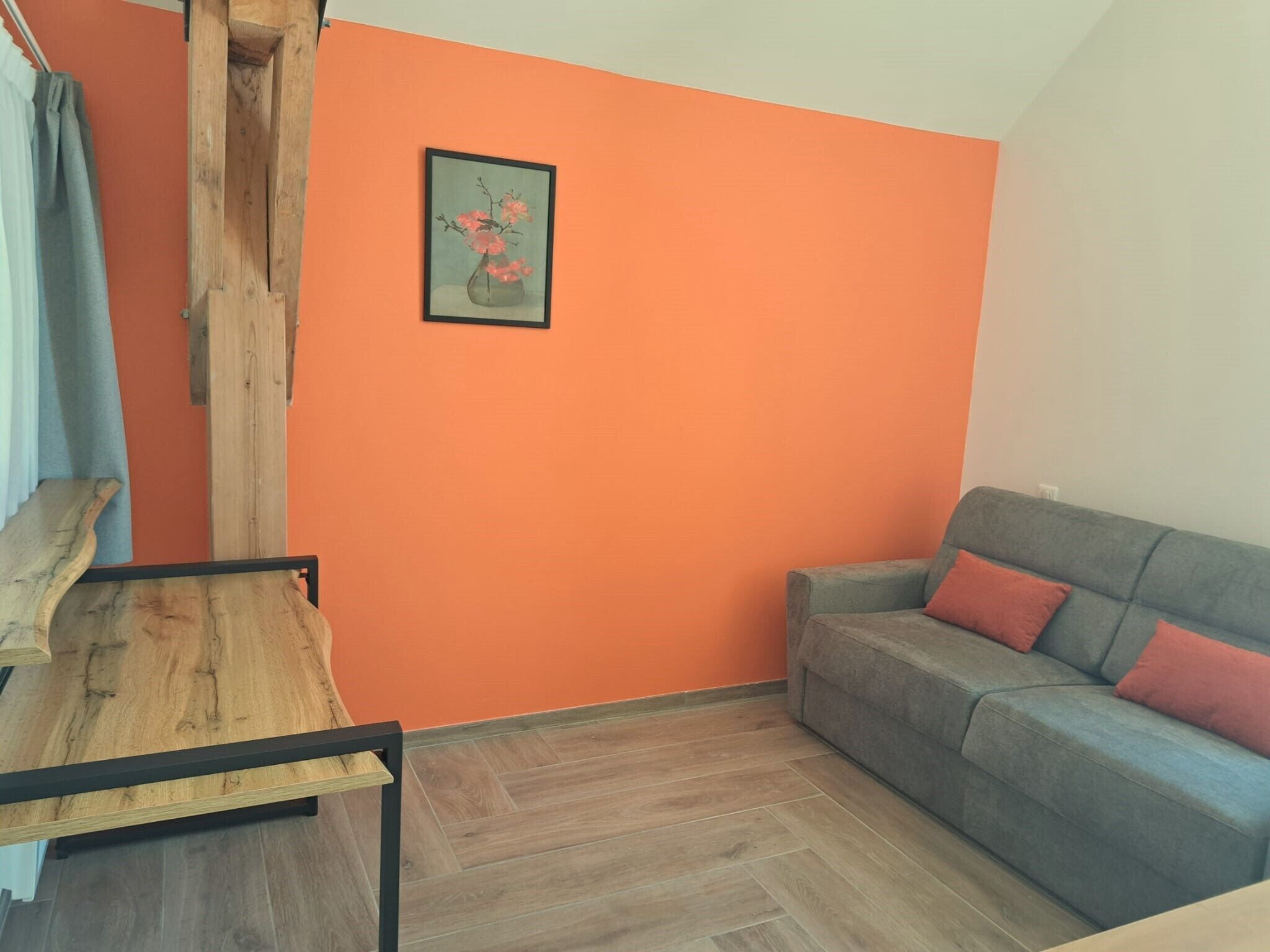 A room with an orange wall, a grey sofa with red cushions, a wooden table, and a framed picture of flowers.